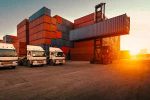 A reach stacker moving shipping containers next to freight trucks, representing complex transportation management operations.