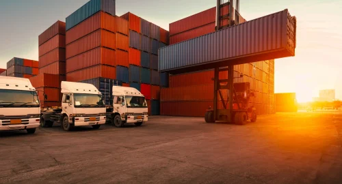 A reach stacker moving shipping containers next to freight trucks, representing complex transportation management operations.